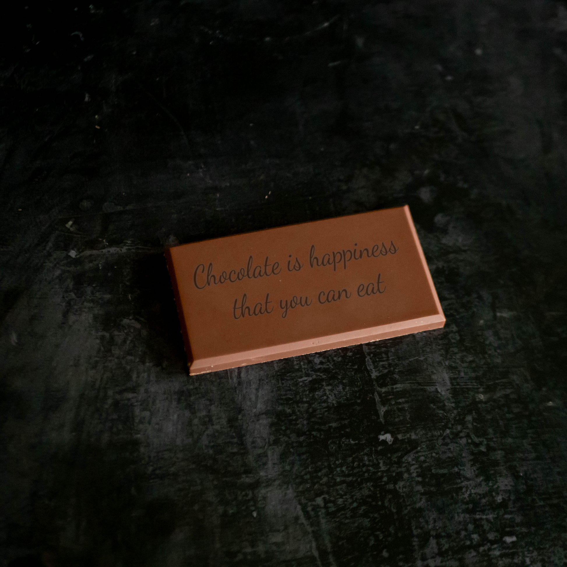 A personalized Belgian milk chocolate bar with text 'Chocolate is happiness that you can eat' printed on it, placed on a dark surface.