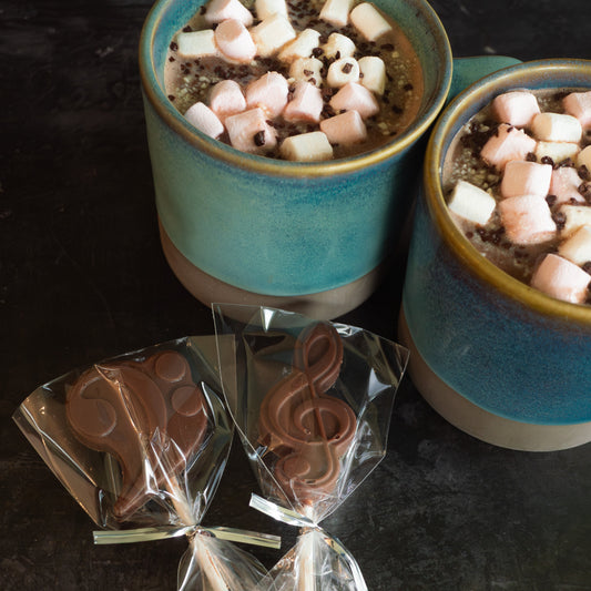 Two mugs of hot chocolate with marshmallows on a dark surface, accompanied by two wrapped chocolate candies.