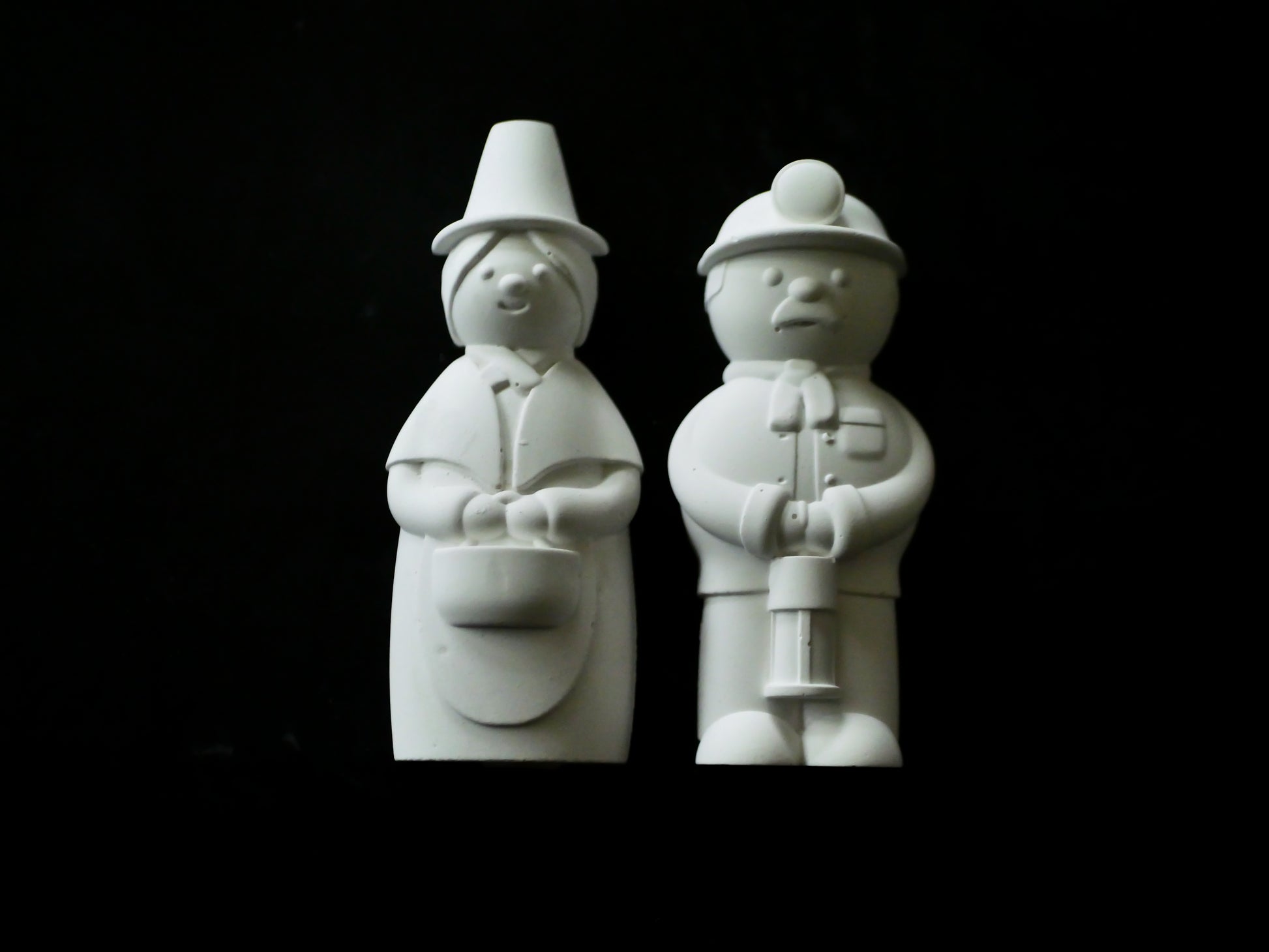 Two white ceramic figures of a Welsh miner and a Welsh ladyon a black background