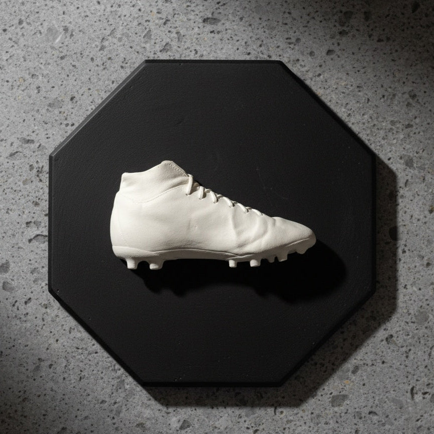 White soccer cleat on a black octagonal platform with a concrete floor background