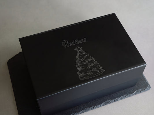 A black rectangular gift box with a glossy finish, featuring an engraved Christmas tree and the brand name 'RedChaos' on the lid, resting on a dark slate coaster against a neutral gray background.