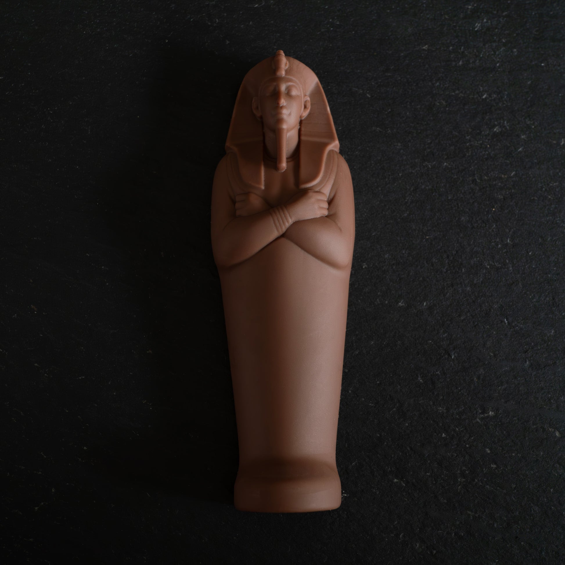 An Egyptian mummy sarcophagus figurine in a chocolate , standing upright with arms crossed, set against a dark textured slate background.
