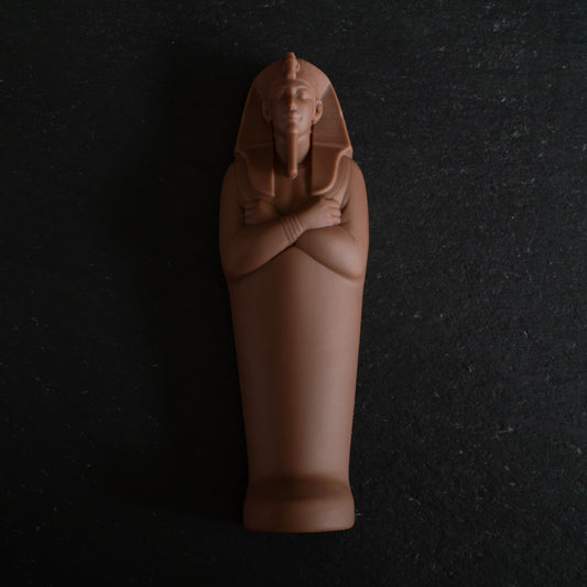 An Egyptian mummy sarcophagus figurine in a chocolate , standing upright with arms crossed, set against a dark textured slate background.