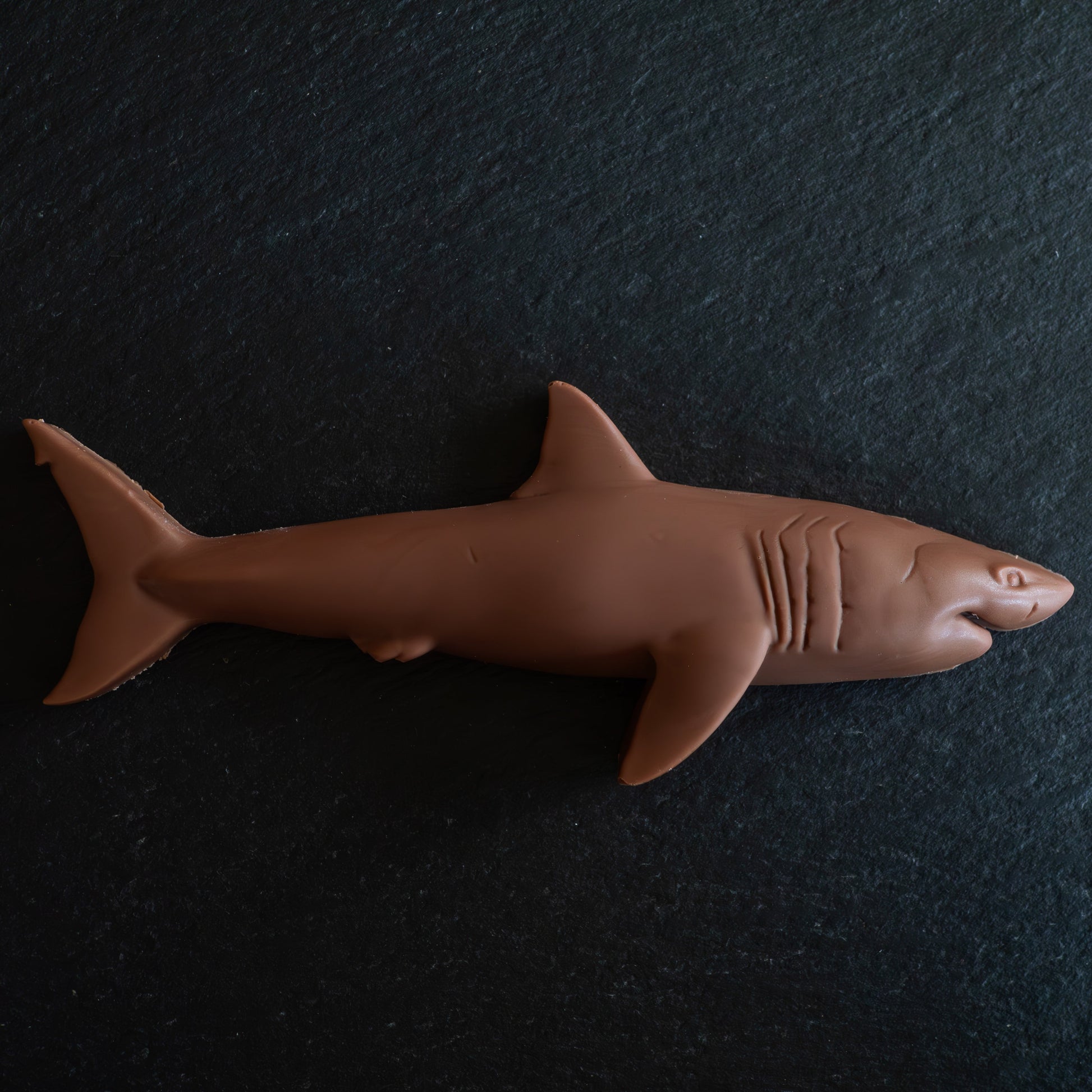 A brown shark-shaped confection, likely a chocolate bar or candy, displayed on a dark textured slate background.